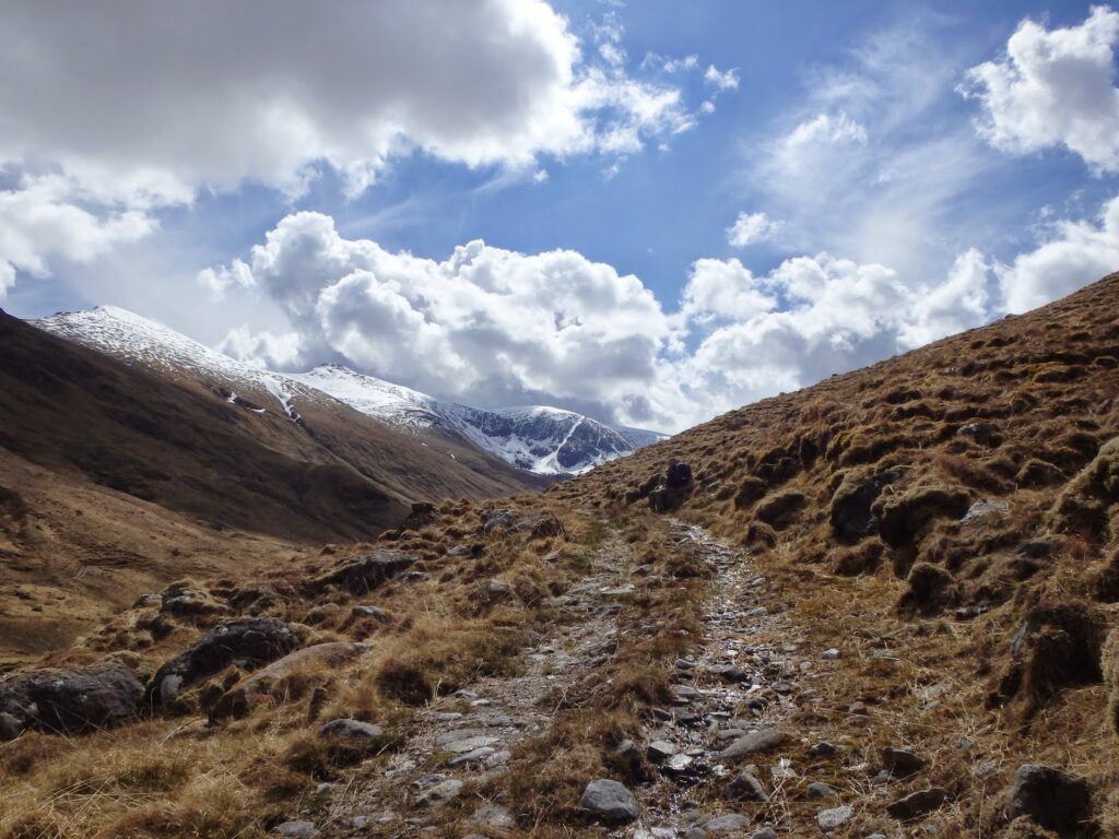 rocky path leading over hillside to snowy mountains. Sky with sun and clouds